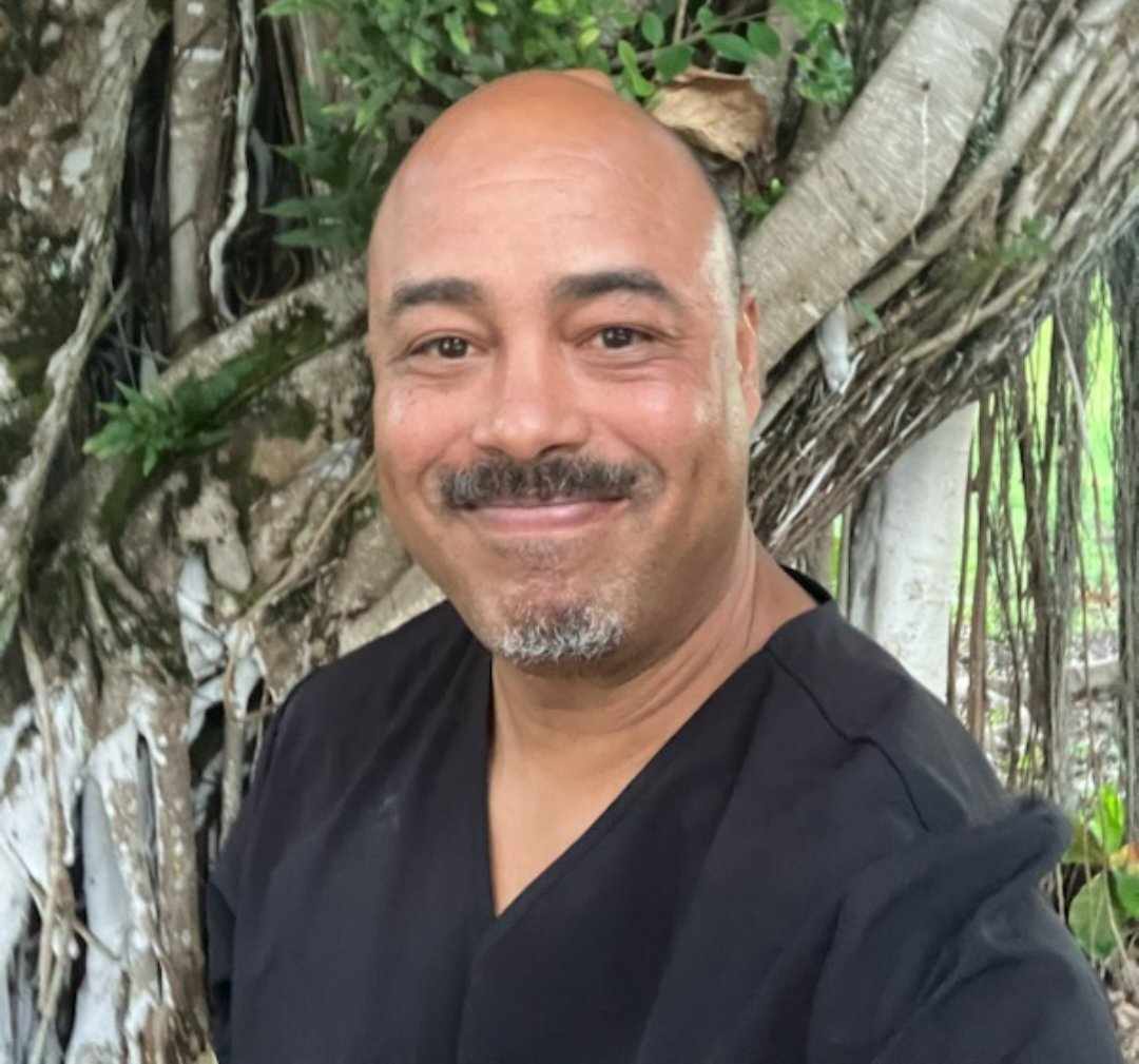 David is wearing a black scrub top in front of a tree with vines.