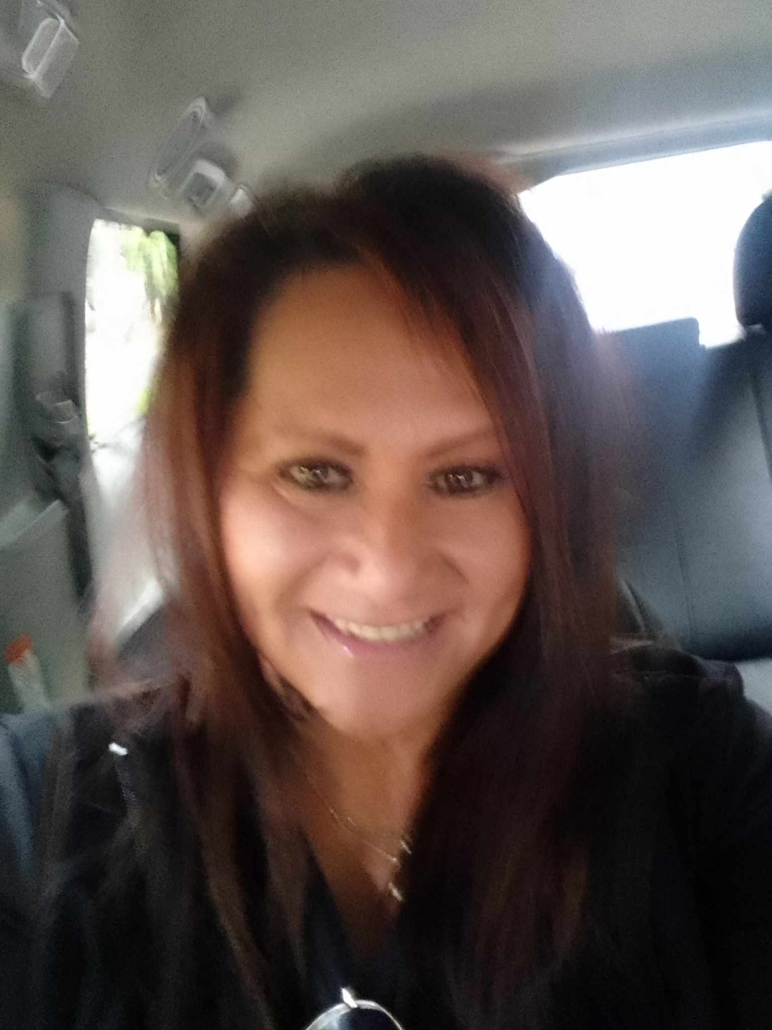Kelani has dark eyeliner and is wearing a dark scrub top sitting in the front seat of a vehicle.