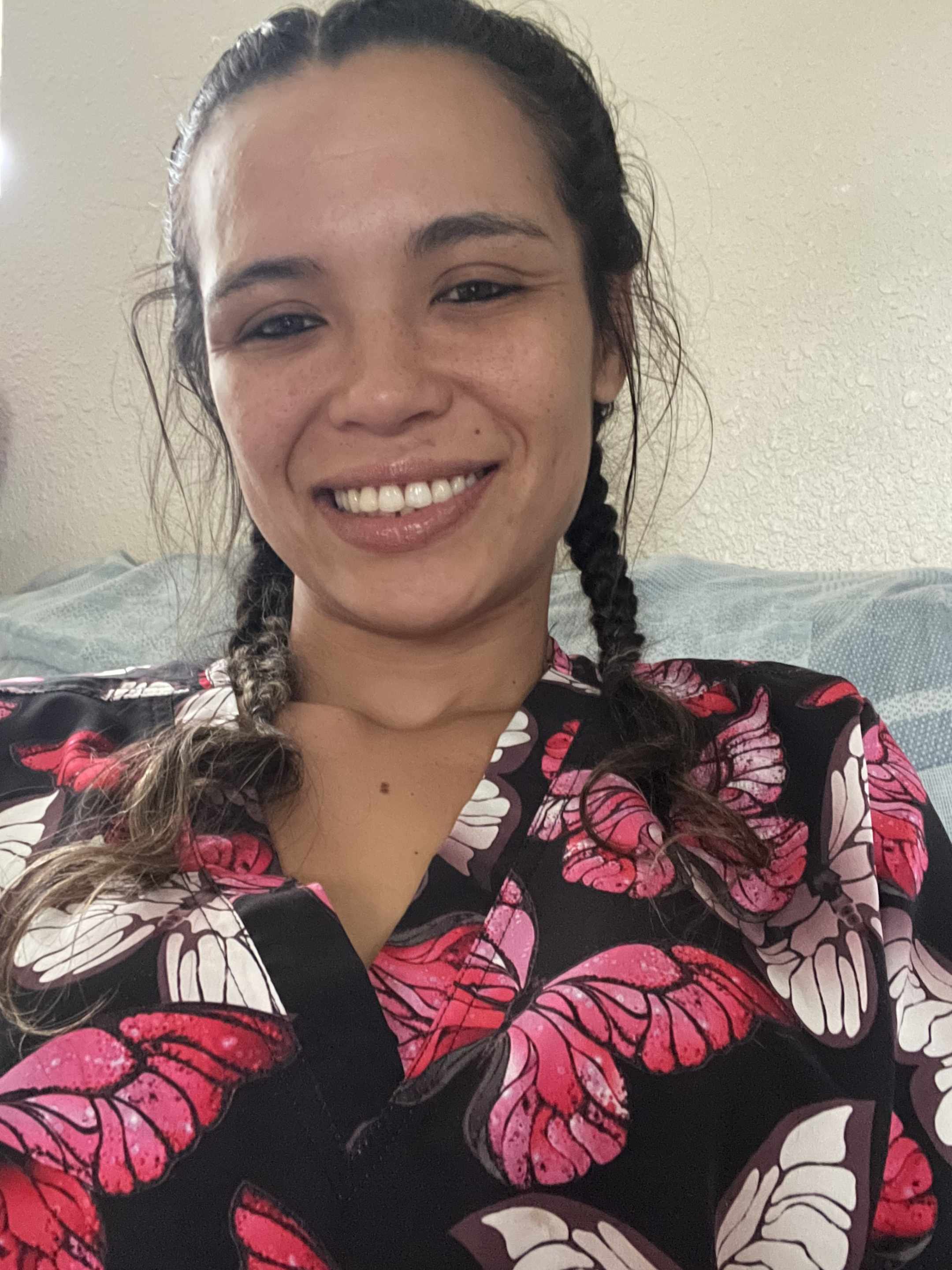 Kiana has dark brown hair in two braids at the sides and is wearing dark scrubs with a pink butterly pattern in front of a white wall.