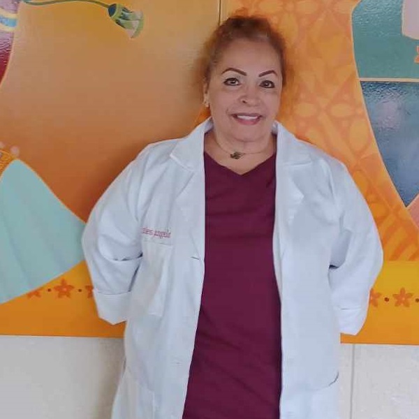 Lita has a medium complexion and is wearing a white lab coat, burgundy scrubs, and makeup. She is standing in front of an orange wall.