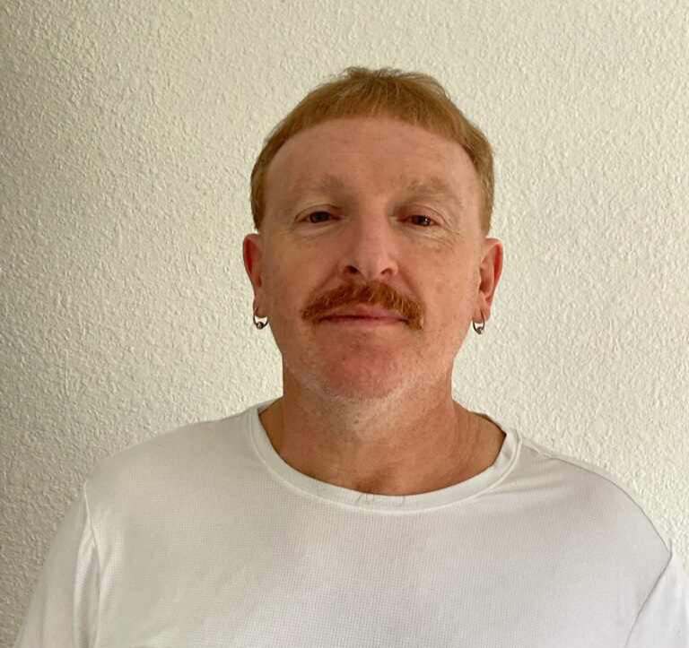 Michael has ginger hair and freckles and wears a plain white tee shirt and golden hoop earrings against an off white wall. He has a ginger mustache.